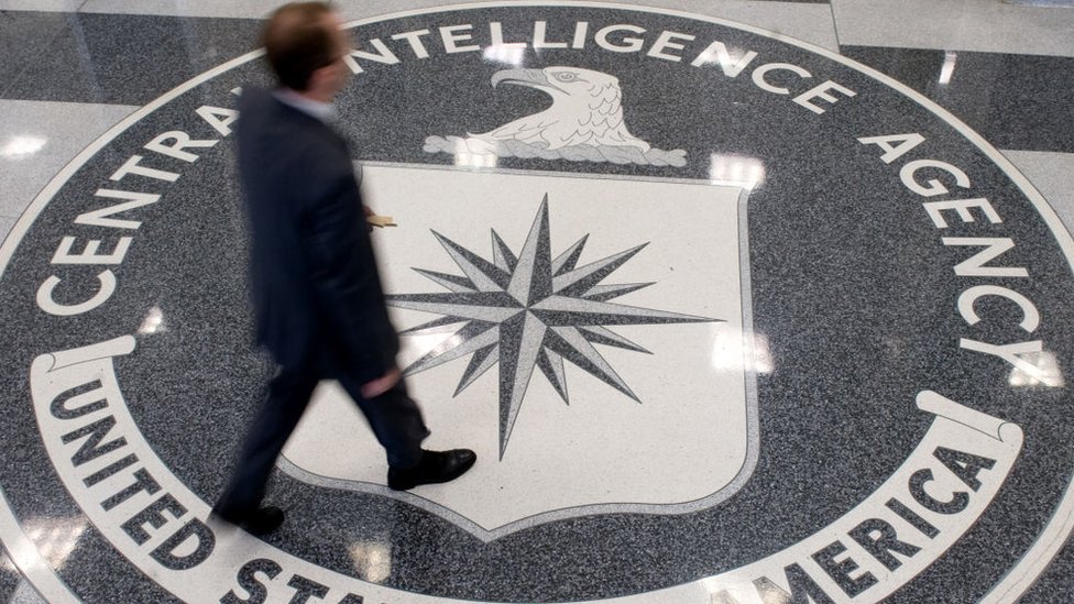 A person in a suit walking across the seal of the Central Intelligence Agency inlaid in a lobby floor.